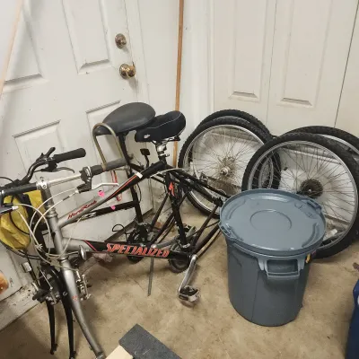 Sale Photo Thumbnail #142: Two Specialized Hardrock mountain bicycle frames stored without front wheels attached, alongside detached bicycle wheel sets and a grey trash bin.