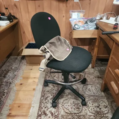 Sale Photo Thumbnail #124: Black fabric armless drafting stool with height adjustment and foot ring, resting next to a wooden desk.