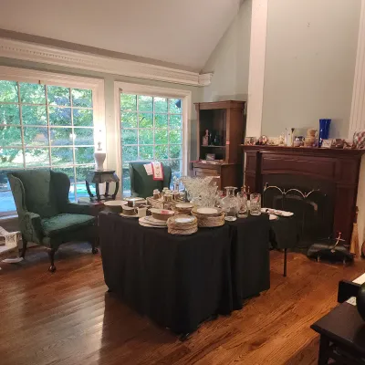 Sale Photo Thumbnail #145: Assorted glassware, china, and crystal displayed on black tablecloths in a living room, featuring green upholstered wingback chairs and a dark wood fireplace mantle in the background.