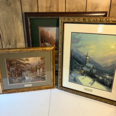Sale Photo Thumbnail #107: Group of three framed Thomas Kinkade art prints featuring picturesque winter and village scenes, housed in decorative molded and gilded wooden frames with double matting.