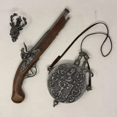 Sale Photo Thumbnail #18: Vintage replica flintlock pistol accompanied by an ornate embossed metal powder flask with a leather strap and a small stag head wall accent.