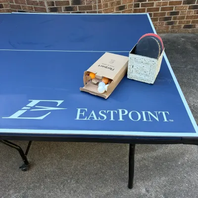 Sale Photo Thumbnail #80: Blue EastPoint ping pong table with a cardboard box of table tennis balls and a vintage-style holder containing paddles.