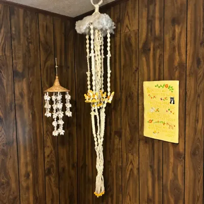 Sale Photo Thumbnail #188: Vintage 1970s macramé plant hanger with carousel figure accents and faux cloud top, displayed alongside a shell wind chime against wood paneling.