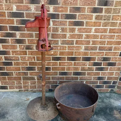 Sale Photo Thumbnail #68: Vintage red cast-iron hand water pump mounted on a stand, paired with a large rustic cast-iron cauldron, showing significant patina and surface rust.