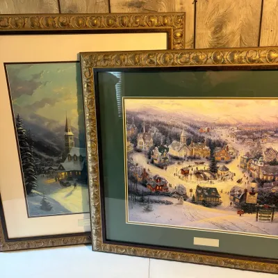 Sale Photo Thumbnail #106: Two framed Thomas Kinkade art prints depicting cozy winter village scenes, housed in ornate metallic gold frames with matting.