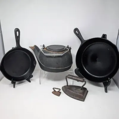 Sale Photo Thumbnail #160: Collection of vintage cast iron cookware and antique items, including two skillets on display stands, a tea kettle with a wire handle, and two sad irons.