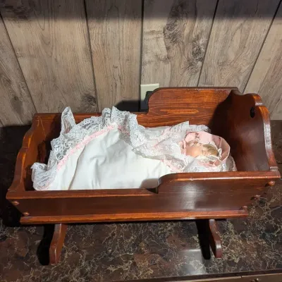 Sale Photo Thumbnail #84: Vintage dark stain wooden doll cradle with rocking runners, featuring a sleeping porcelain baby doll dressed in a lace-trimmed bonnet and matching blanket.