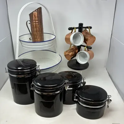 Sale Photo Thumbnail #169: Collection of modern kitchenware featuring a set of four black ceramic canister jars with clamp lids, copper mugs with white linings on a black stand, and a white enamel two-tiered wire rack with copper pitcher.