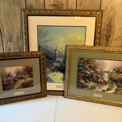 Sale Photo Thumbnail #141: Trio of framed, matted Thomas Kinkade art prints housed in ornate gilded frames, featuring cozy landscape and village scenes.