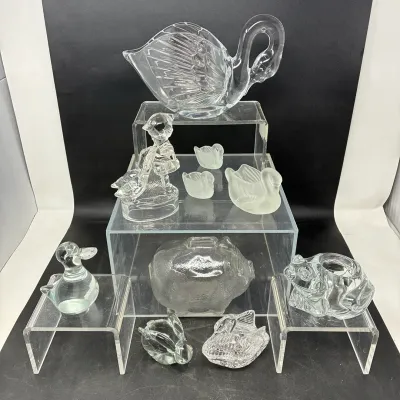 Sale Photo Thumbnail #163: Collection of clear and frosted glass figurines and decorative items, featuring various swans, a pig coin bank, a frog candle holder, and a duck, displayed on tiered acrylic stands.