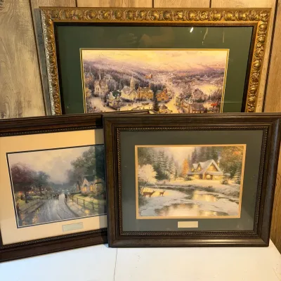 Sale Photo Thumbnail #108: Trio of framed Thomas Kinkade art prints, including a large landscape in an ornate gold-tone frame and two smaller cottage scenes in dark wood frames.