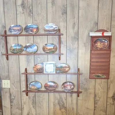 Sale Photo Thumbnail #55: Collection of oval decorative plates depicting rural scenes displayed on wall-mounted wooden shelves, with an additional wooden rack to the right.
