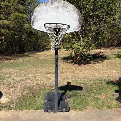 Sale Photo Thumbnail #69: Portable outdoor basketball hoop with a fillable plastic base, metal pole, net, and a weathered fan-shaped backboard.