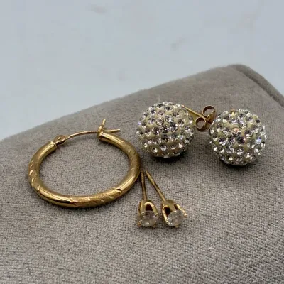 Sale Photo Thumbnail #200: Assortment of gold-tone pierced earrings, including a single textured hoop, crystal disco ball studs, and small solitaire stud earrings.