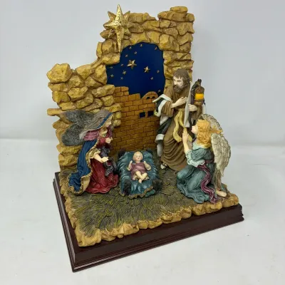 Sale Photo Thumbnail #180: Detailed resin Nativity scene diorama on a dark wooden base, featuring Mary, Joseph, Baby Jesus, and a kneeling angel set against a stone stable wall under a starry night sky.