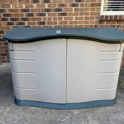 Sale Photo Thumbnail #78: Large outdoor resin storage cabinet with a dark green contoured top lid and light gray double front doors, placed against a brick wall.