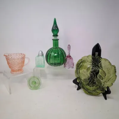 Sale Photo Thumbnail #156: Assorted vintage glass collectibles, including a green decanter, pink depression glass creamer, small pink glass bell, salt shaker, and olive green embossed plate displayed on risers and a stand.