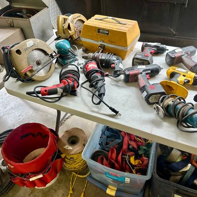 Sale Photo Thumbnail #17: Assortment of power tools, including circular saws, drills, impact drivers, and angle grinders, displayed on a folding table alongside storage tubs of straps and roping.