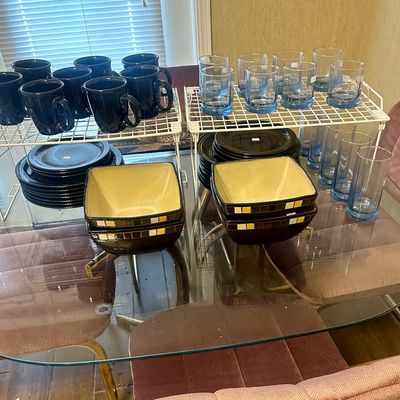 Sale Photo Thumbnail #58: Collection of contemporary kitchenware, including dark ceramic mugs and dinner plates, square serving bowls with geometric trim, and light blue glassware set on a glass-top table.