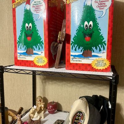 Sale Photo Thumbnail #49: Two boxed Douglas Fir The Talking Tree vintage animated Christmas decorations resting on a wire shelf above small dog figurines, a baseball cap, and novelty decor.
