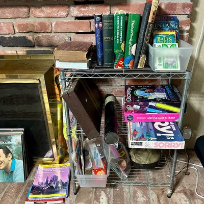 Sale Photo Thumbnail #32: A small wire shelving unit holding vintage hardcovers, board games, and craft kits next to a brick fireplace.