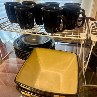 Sale Photo Thumbnail #59: Collection of black ceramic dinnerware including mugs and plates, paired with two square beige and brown serving bowls displayed on a wire rack.