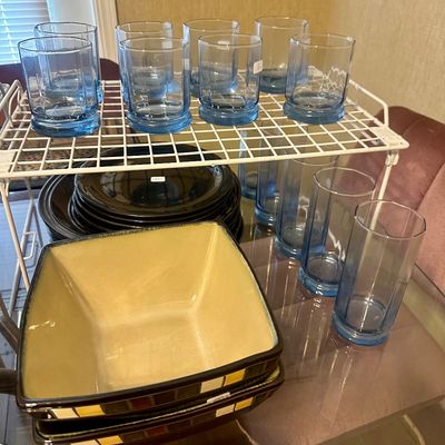 Sale Photo Thumbnail #60: Collection of blue tint drinking glasses, black ceramic plates, and square yellow ceramic serving bowls organized on a glass tabletop and wire rack.