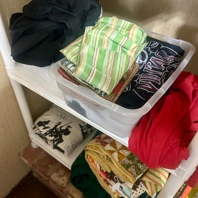 Sale Photo Thumbnail #48: Assorted folded textiles and clothing arranged on a white plastic shelving unit, including a clear plastic storage bin, patterned quilts, and various colored garments.