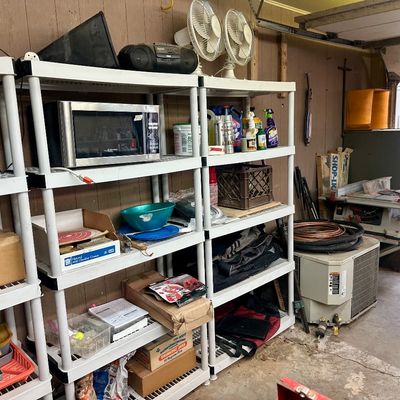 Sale Photo Thumbnail #26: Garage storage area filled with plastic shelving units holding household electronics, small appliances, cleaning supplies, tools, and a table saw.
