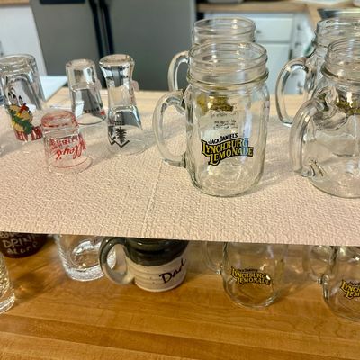 Sale Photo Thumbnail #61: Collection of novelty glassware, including Jack Daniel's Lynchburg Lemonade mason jar mugs and assorted souvenir shot glasses, displayed on a wire shelf.