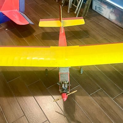 Vintage gas-powered remote control airplane with a yellow and red monoplane design, fitted with a front propeller and landing gear.
