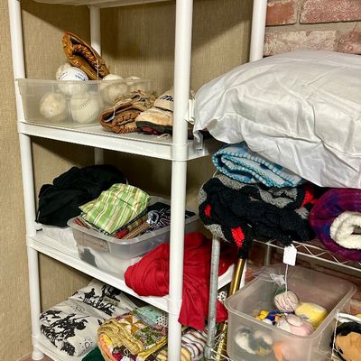 Sale Photo Thumbnail #35: White plastic four-tier shelving unit loaded with household goods, including baseball equipment, folded linens, quilts, a duffel bag, and a yellow top hat.