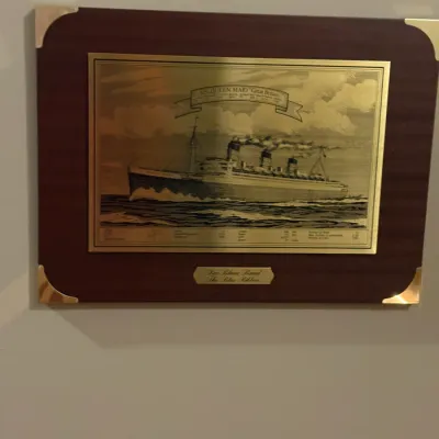 Sale Photo Thumbnail #82: Etched brass plaque mounted on wood depicting the ocean liner R.M.S. Queen Mary, titled 'The Blue Ribbon'.