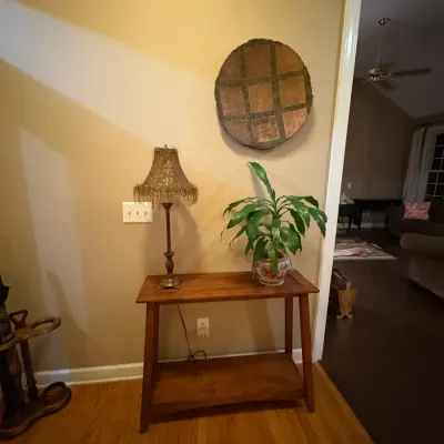 Sale Photo Thumbnail #22: Wooden two-tier console table holding a table lamp with a fringed shade and a potted houseplant, 