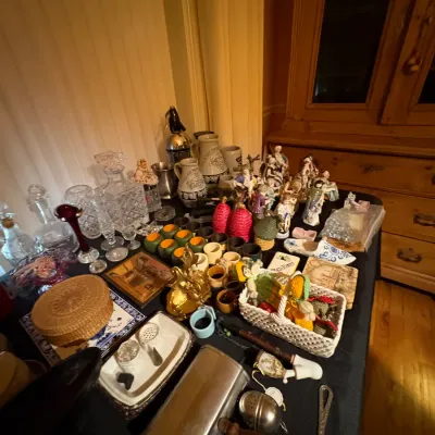 Sale Photo Thumbnail #21: A tabletop filled with vintage collectibles, featuring cut glass decanters, ceramic beer steins, figurines, small baskets, and household knick-knacks.