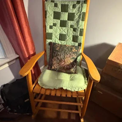 Sale Photo Thumbnail #79: High-back wooden rocking chair with light oak finish, dressed with a green geometric quilt, ruffled seat cushion, and floral accent pillow.