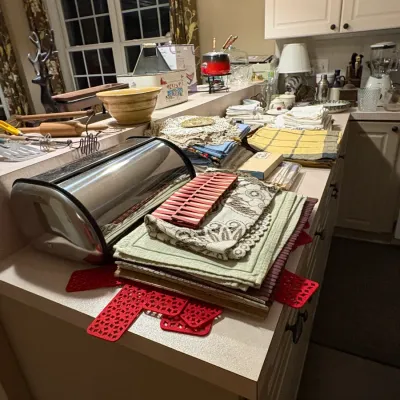 Sale Photo Thumbnail #37: Collection of kitchenware and linens arranged on a kitchen counter, including a stainless steel roll-top bread box, placemats, napkins, a ceramic mixing bowl, and utensils.