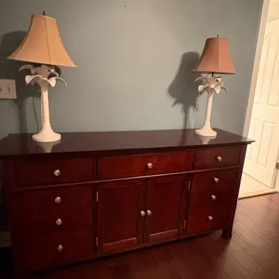Sale Photo Thumbnail #56: Dark mahogany-finish wooden sideboard with multiple drawers and center cabinets, topped with a pair of white palm tree-style table lamps.