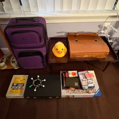 Sale Photo Thumbnail #80: Collection of miscellaneous items including a purple wheeled suitcase, a yellow rubber duck, a leather briefcase, an organic chemistry molecular model set, and various board games.