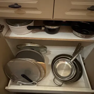 Sale Photo Thumbnail #40: Kitchen cabinet filled with cookware, including stainless steel frying pans, glass pot lids in an organizer, a white ceramic casserole dish, and saucepans.