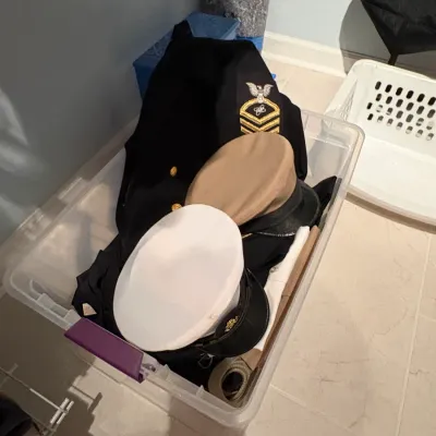 Sale Photo Thumbnail #59: Clear plastic storage tote filled with U.S. Navy dress uniforms, including a black jacket with Master Chief Petty Officer insignia and combination cover hats.