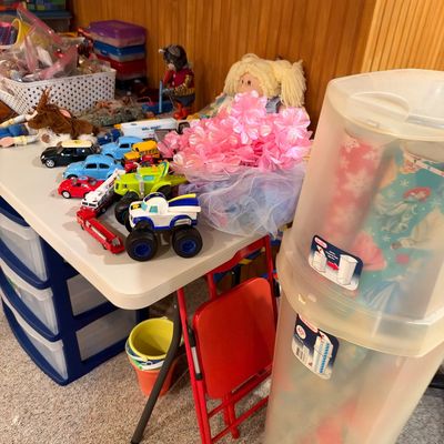 Sale Photo Thumbnail #114: Assortment of children's toys on a folding table, including toy cars, stuffed animals, a doll, pink leis, and a tall plastic wrapping paper storage container.