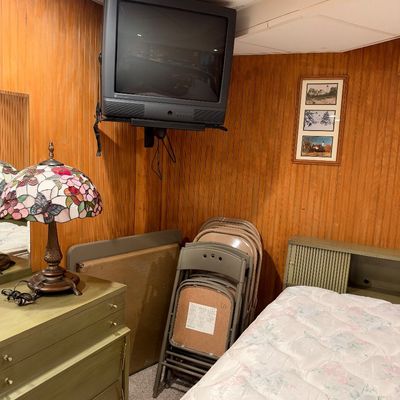 Sale Photo Thumbnail #95: Vintage bedroom setup featuring a ceiling-mounted CRT television, a butterfly motif Tiffany-style lamp, painted wooden dresser, stacked folding chairs, and a twin bed frame.