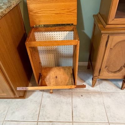 Sale Photo Thumbnail #3: Vintage oak tilt-out trash tilt-bin cabinet with pegboard interior backing, showing moderate wear and detached front trim piece.