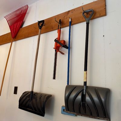Sale Photo Thumbnail #15: Collection of wall-mounted lawn and garden tools, including two snow shovels, an orange hedge trimmer, a long blue-handled tool, and a red leaf rake.