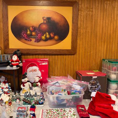 Sale Photo Thumbnail #104: Collection of holiday decorations, including Christmas figurines, ornaments in plastic storage containers, boxed Santa decor, and Santa hats, set below a framed still-life artwork.