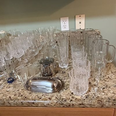 Sale Photo Thumbnail #90: Large collection of crystal and glass barware, including wine stems, highball glasses, beer mugs, and covered metal serving pieces arranged on a granite countertop.