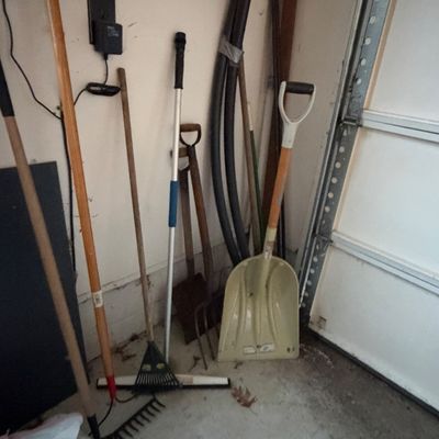Sale Photo Thumbnail #7: Collection of assorted yard and hand tools, including rakes, shovels, a pitchfork, and foam pipe insulation, leaning in a garage corner.