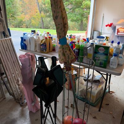 Sale Photo Thumbnail #18: Garage sale setup featuring folding camp chairs, a patio umbrella in a stand, and folding tables filled with various household cleaning supplies and chemicals.