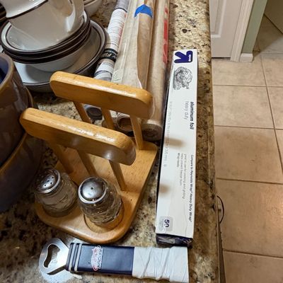 Sale Photo Thumbnail #83: Collection of kitchen items including a wooden salt and pepper caddy, glass shakers, a bottle opener, aluminum foil, and rolls of parchment paper arranged on a granite countertop.